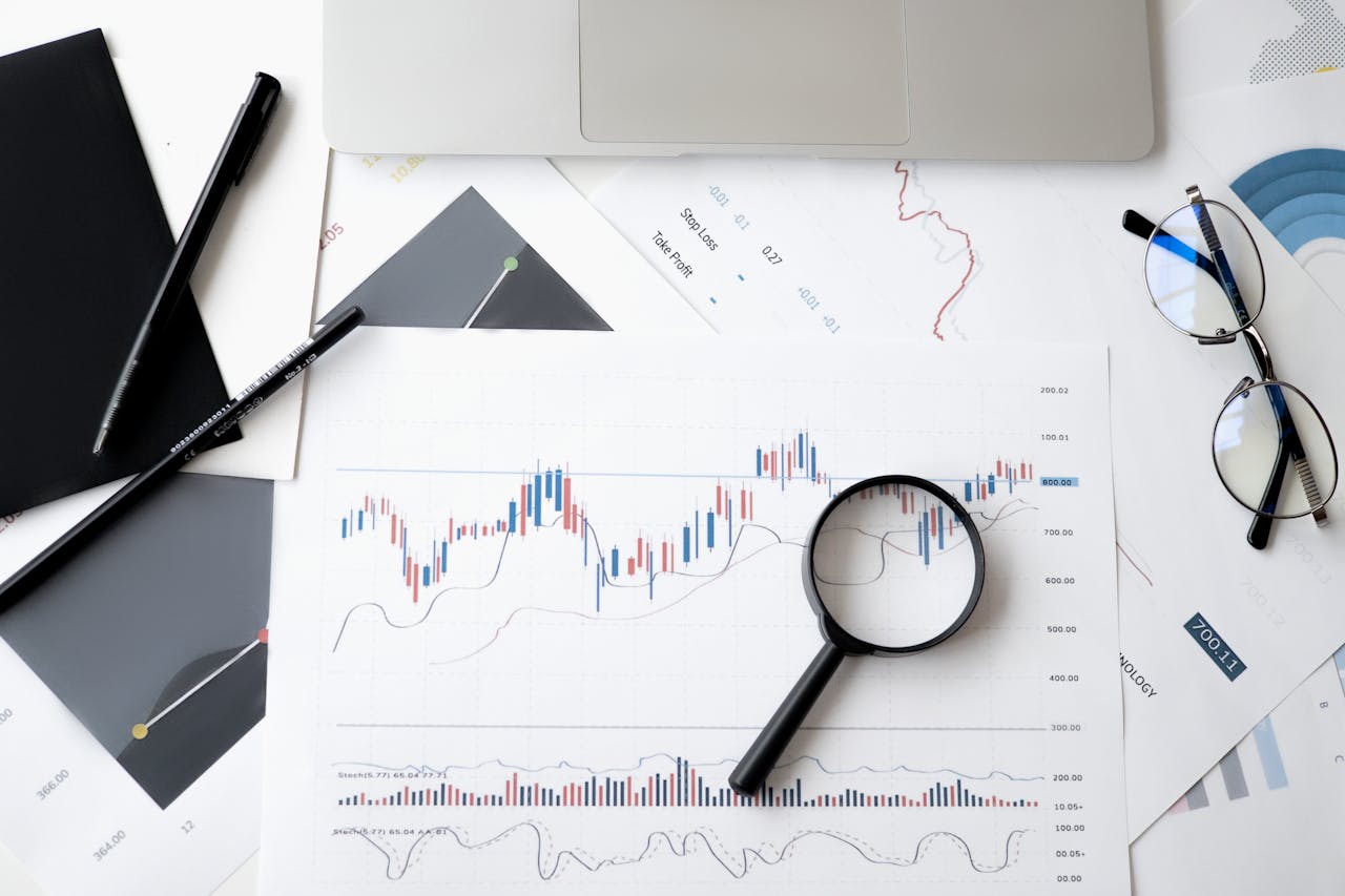 Flat lay of stock market analysis documents with magnifying glass, pens, and glasses.